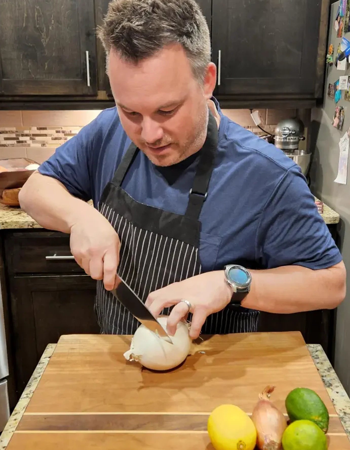 Man in a kitchen cutting an onion on a wooden cutting board with various fruits and vegetables around.