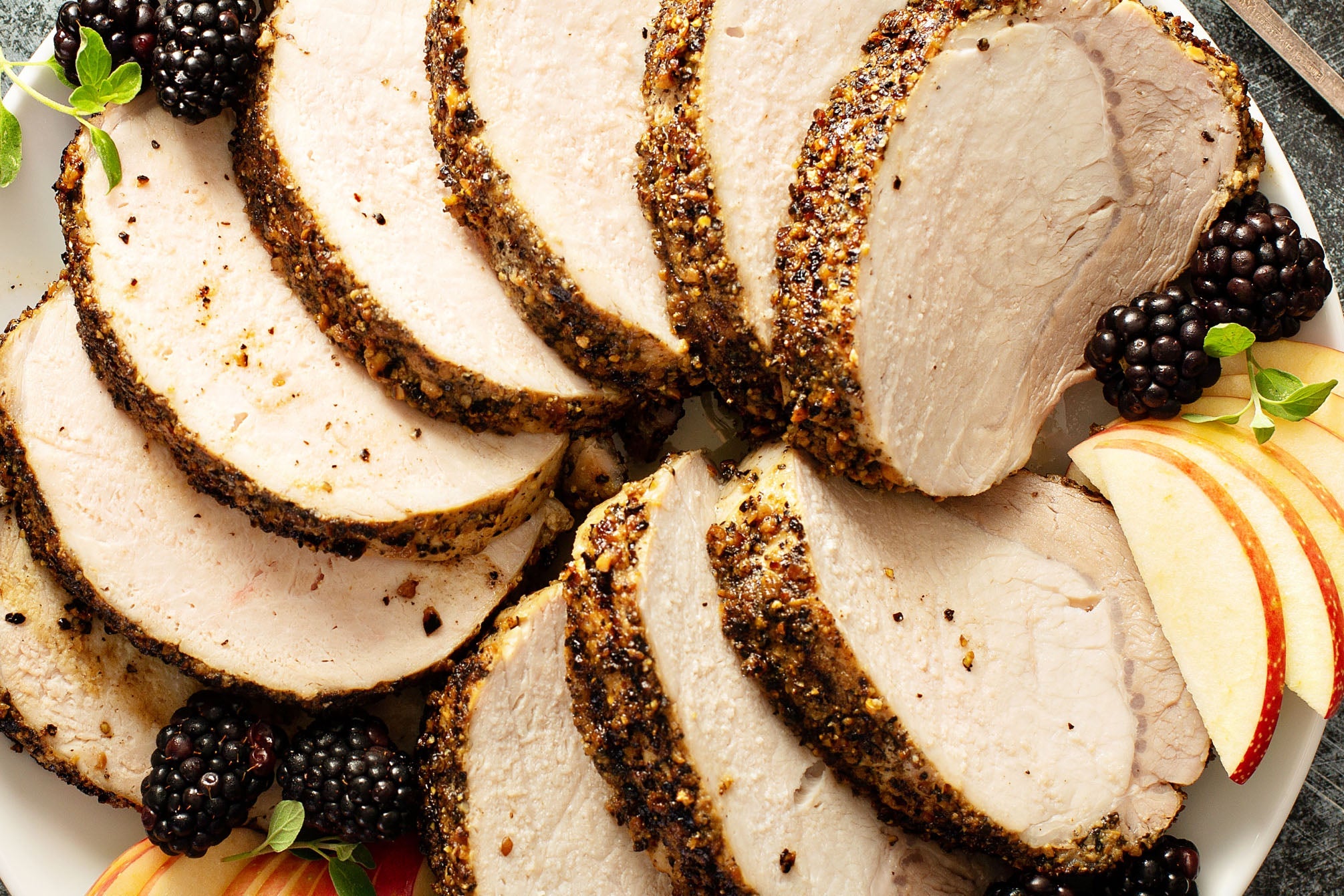 Sliced roasted pork loin on a plate with apples, blackberries, and herbs